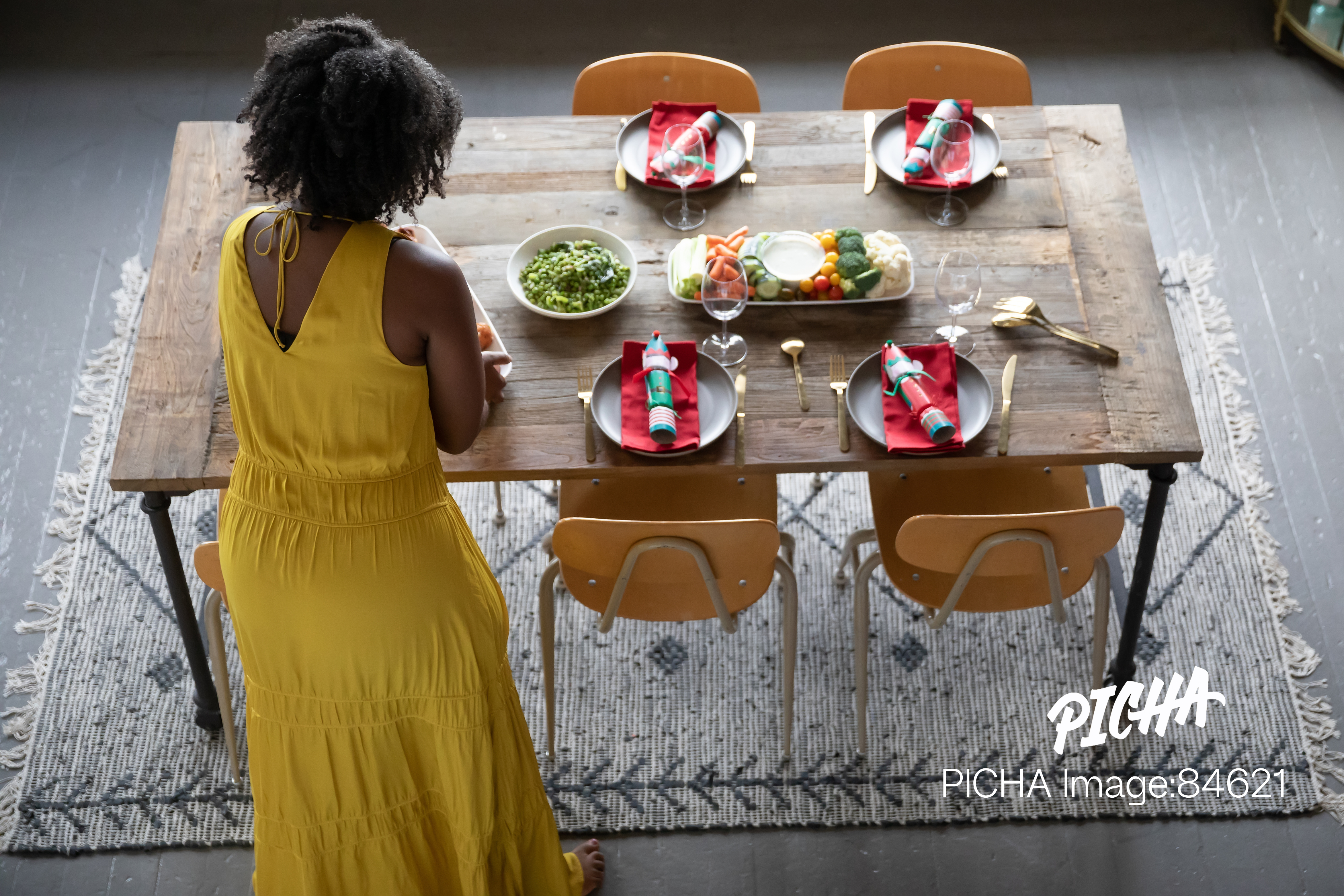 High-angle view of a Black woman in a long yellow dress preparing a rustic wooden dining table for a meal, arranging plates and serving dishes for guests.