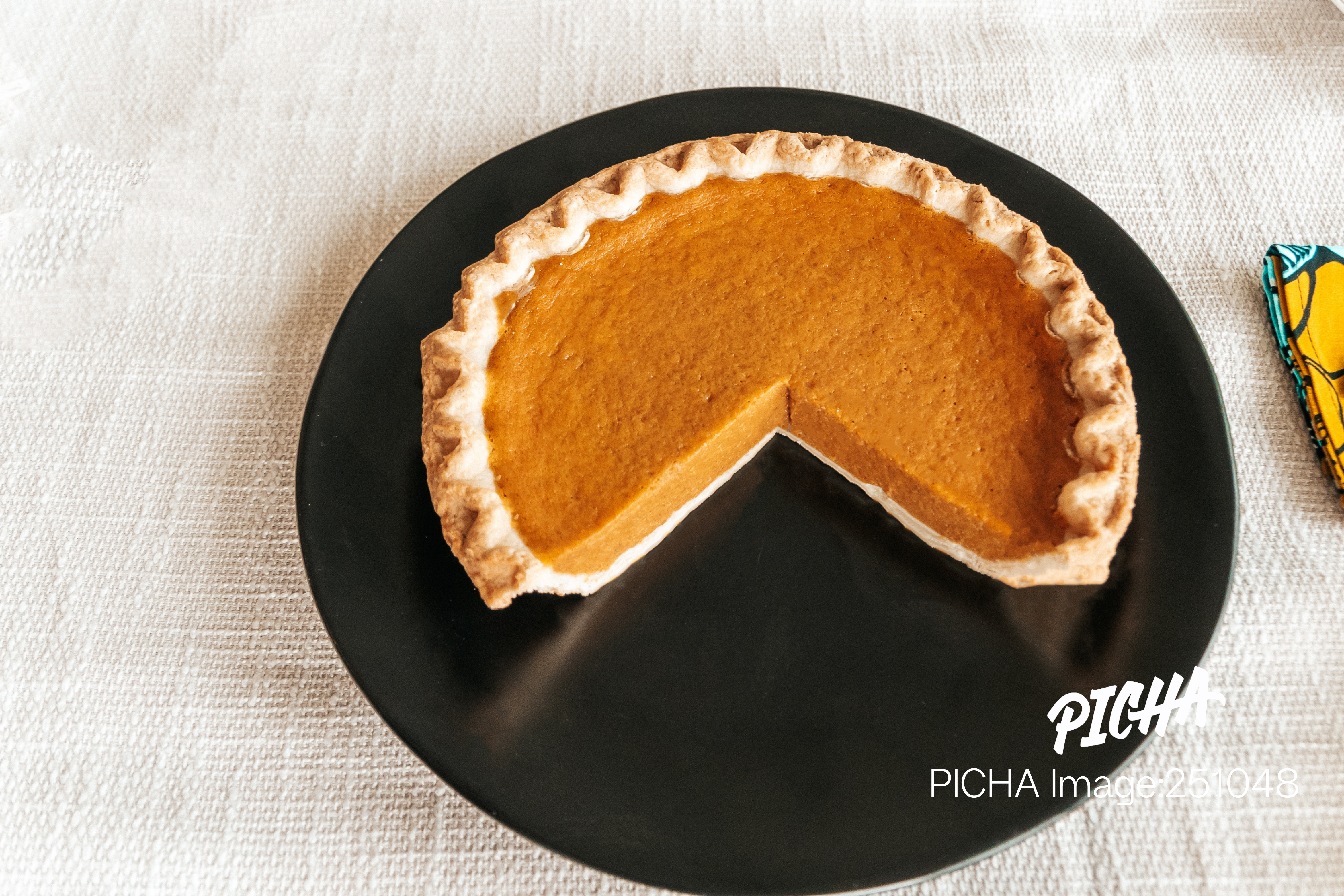 A delicious-looking pumpkin pie with a fluted crust and one slice removed, served on a modern matte black plate on a light-colored tablecloth.