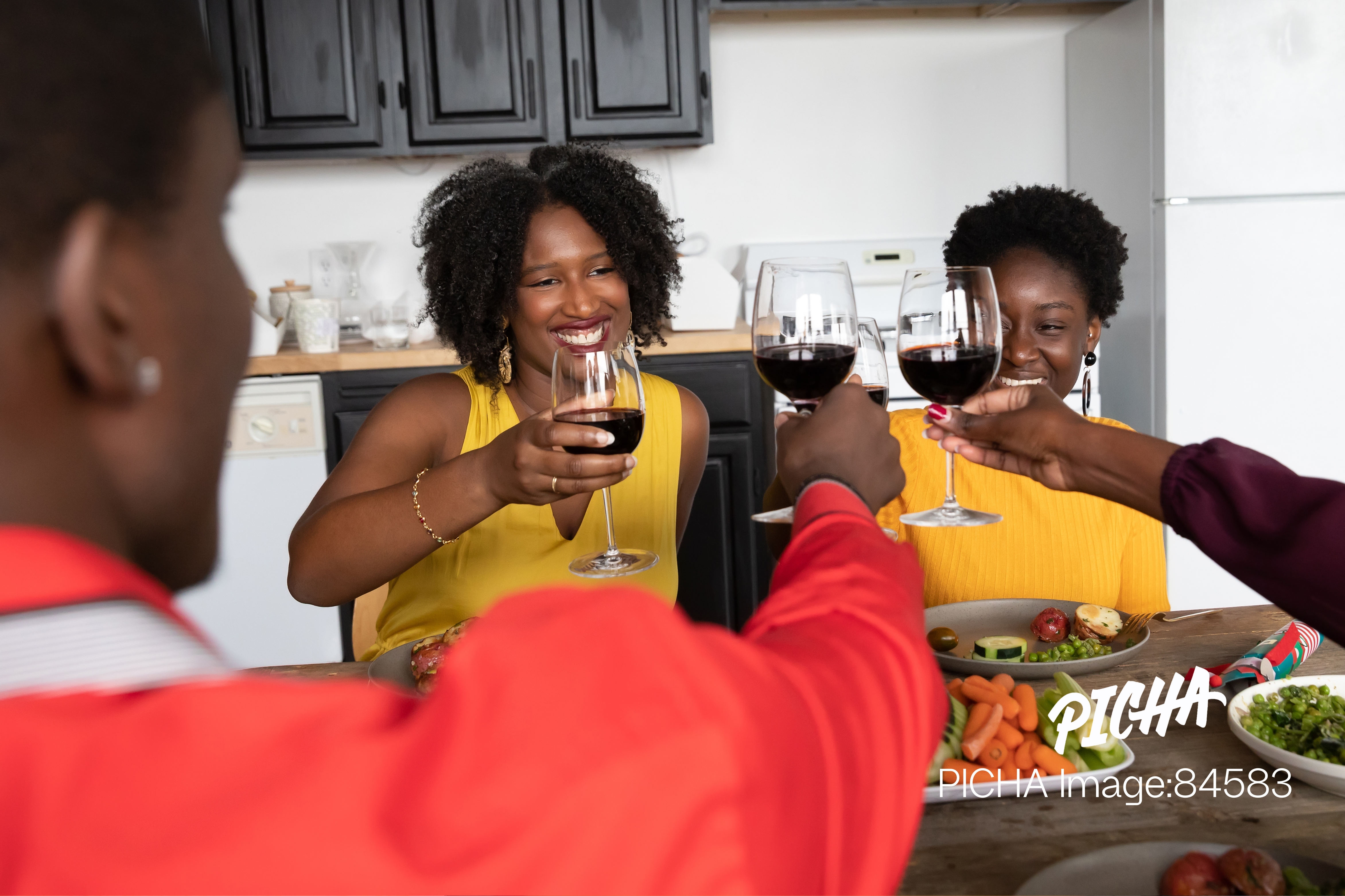 A group of smiling Black friends sitting around a dinner table, raising their glasses of red wine for a celebratory toast in a modern kitchen.