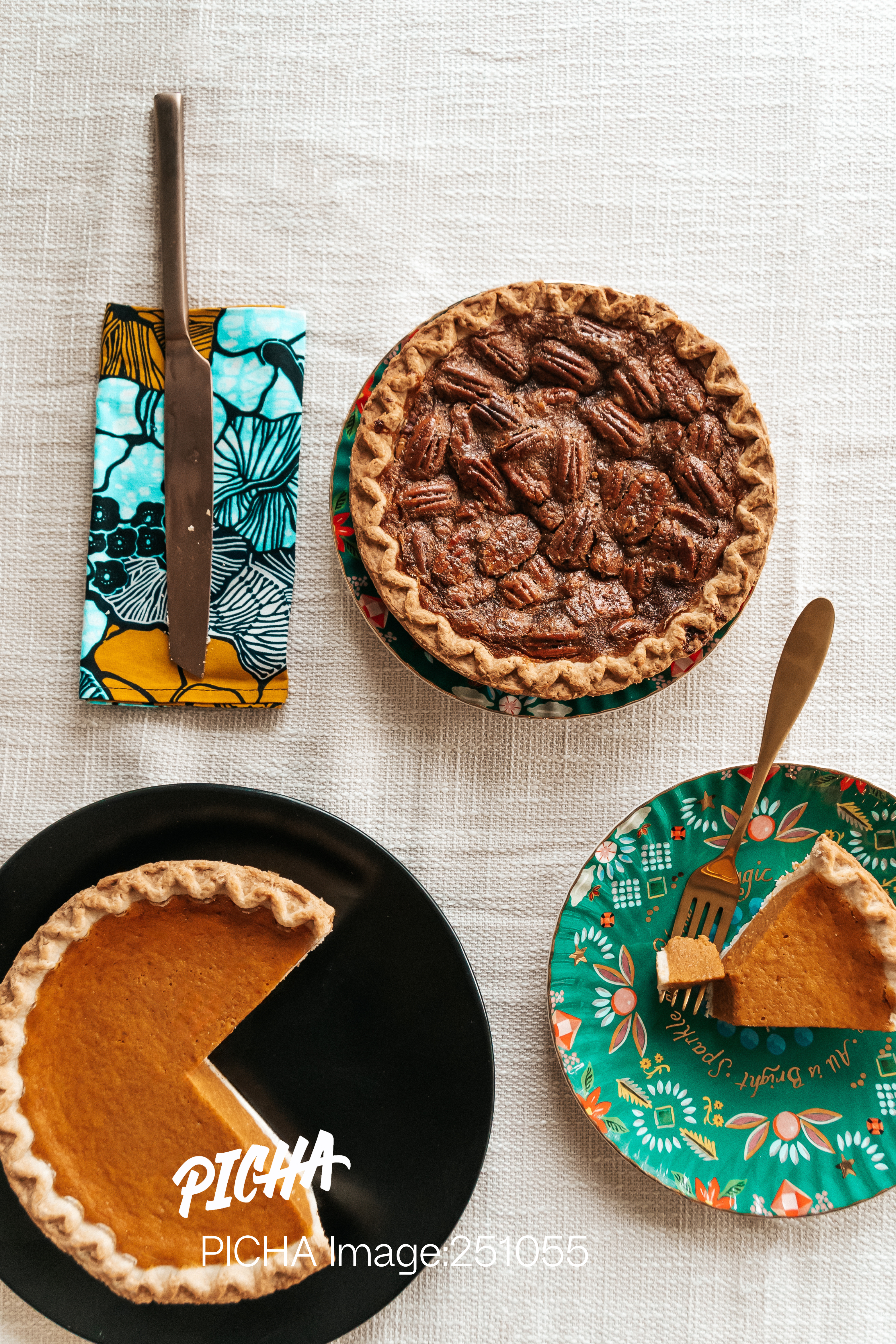 Overhead flat lay view of a holiday dessert table featuring a whole pecan pie and two slices of pumpkin pie served on decorative plates. A knife rests on a colorful blue and yellow African print napkin, adding a cultural touch to the festive scene.