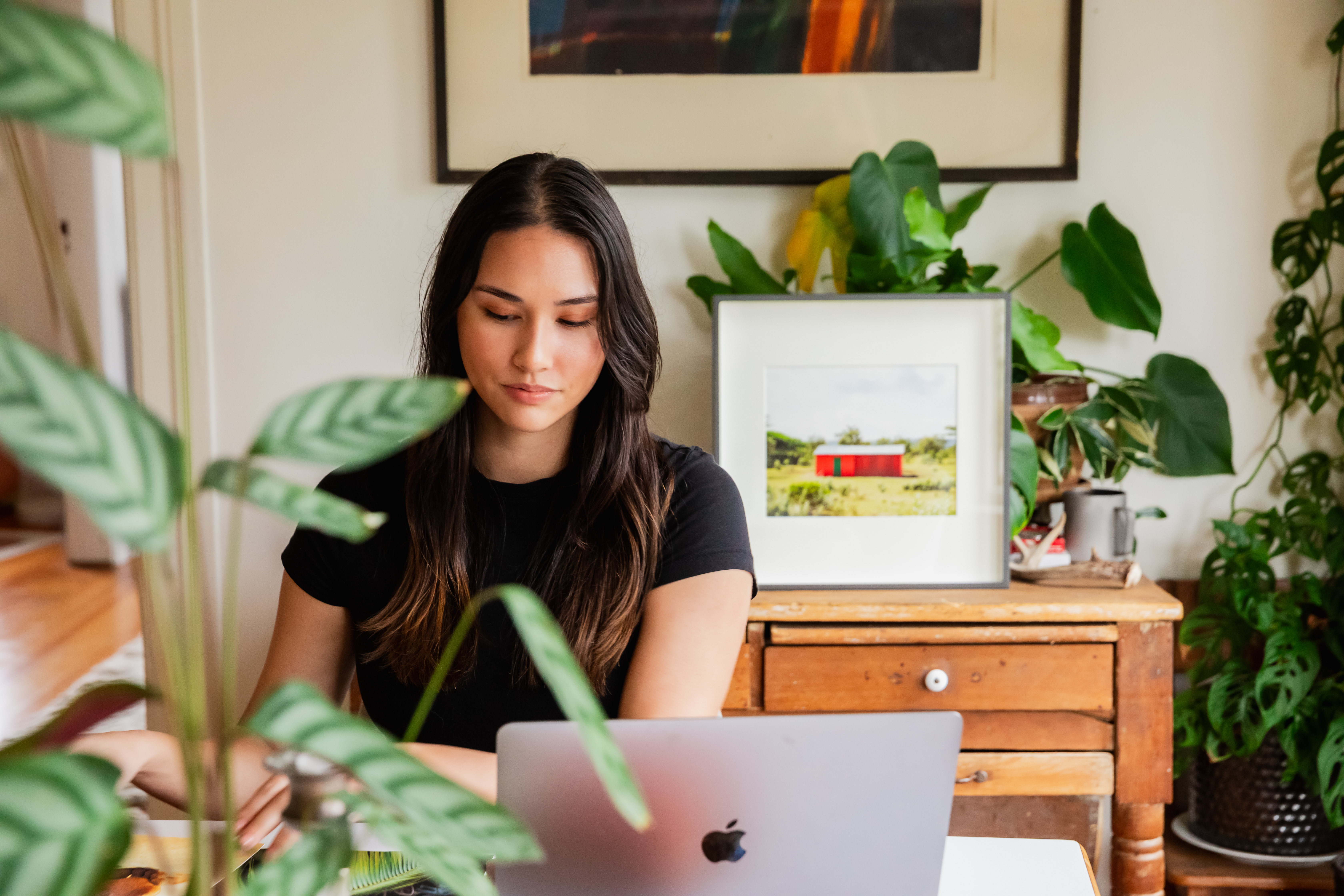 Woman working from home surrounded by plants, with a framed fine art photography print from Rhythm & Hues featuring a red hut in a lush green landscape.